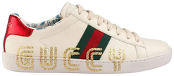 Gucci Wmns Ace Low sneaker in cream leather with “Guccy” side print and metallic heel detail.